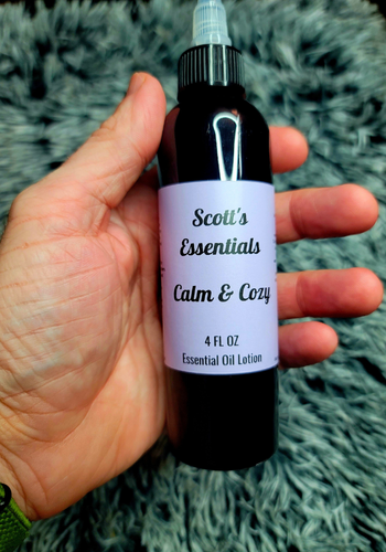 Bottle of 'Scott's Essentials Calm & Cozy' essential oil lotion held in a hand against a textured gray background.