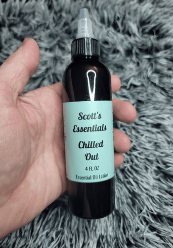 Hand holding a bottle of 'Scott's Essentials Chilled Out' essential oil lotion on a textured gray surface.