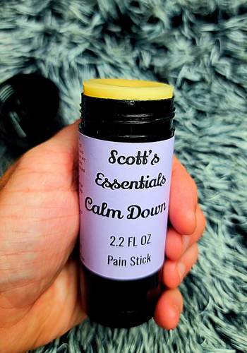 Hand holding a 'Scott's Essentials Calm Down' pain stick against a textured gray background