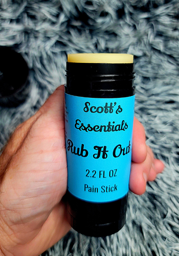 Hand holding a pain stick labeled 'Scott's Essentials Rub It Out' against a textured gray background